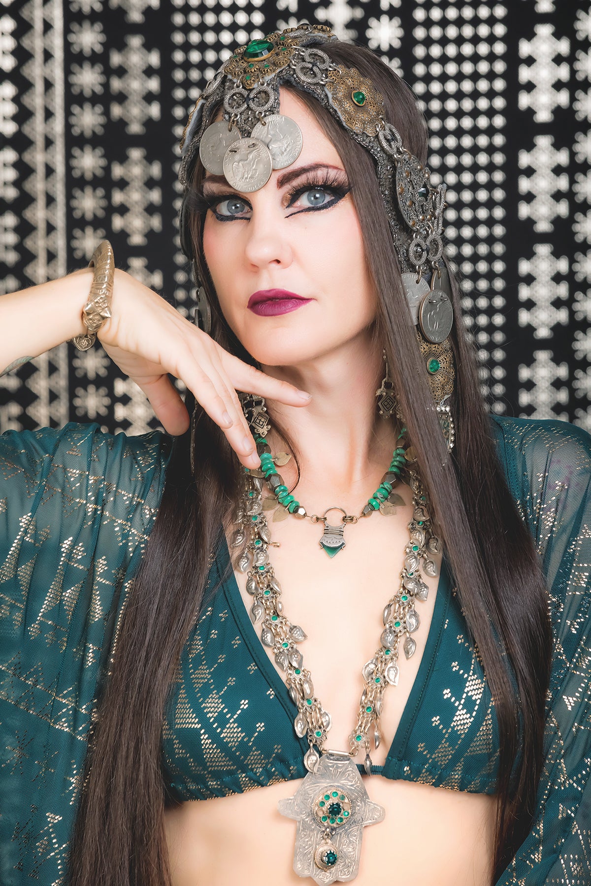 A woman in elaborate Middle Eastern-inspired attire poses gracefully. Her look evokes elegance, standing out like the Alkimiya Bikini Set Malachite by Melodia Designs against a patterned black-and-white background.