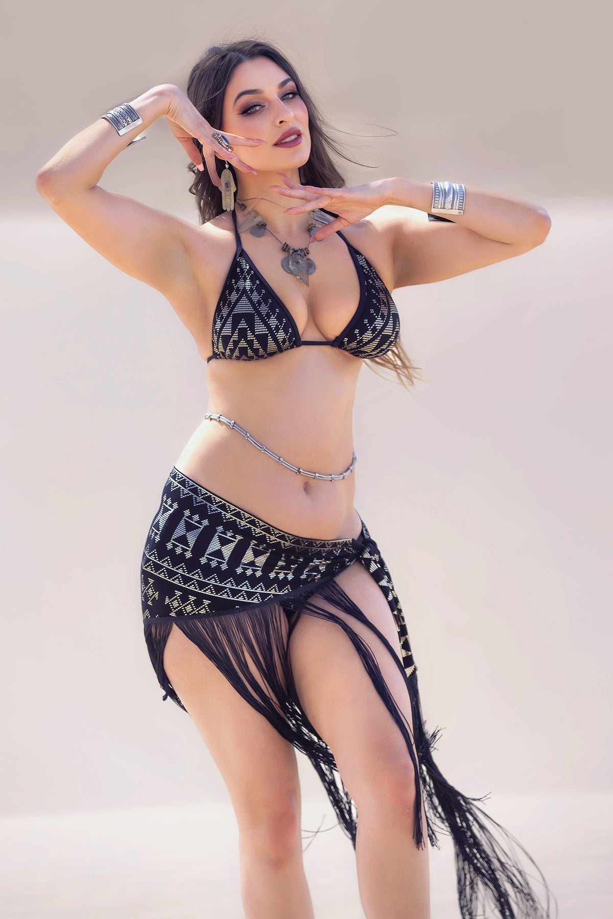 A woman poses outdoors in the Alkimiya Fringe Shimmy Shawl Obsidian by Melodia Designs, styled as a chic resort look. She pairs it with silver jewelry and loose hair, set against a neutral background.