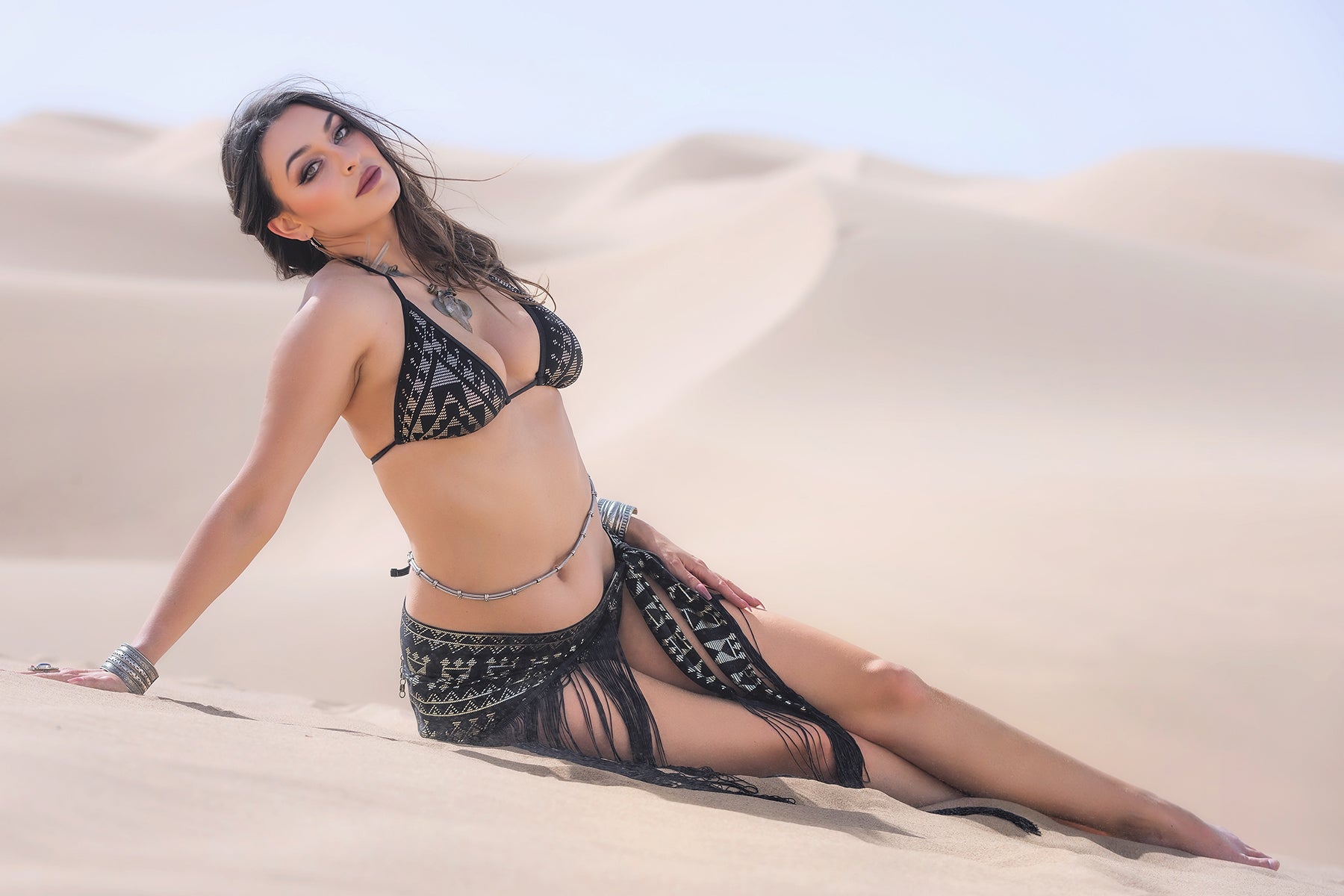 A woman wears the Melodia Designs Alkimiya Lexi Halter Bra Obsidian, sitting on light sand dunes under a clear sky. She looks at the camera, accessorized with jewelry and long wavy hair.