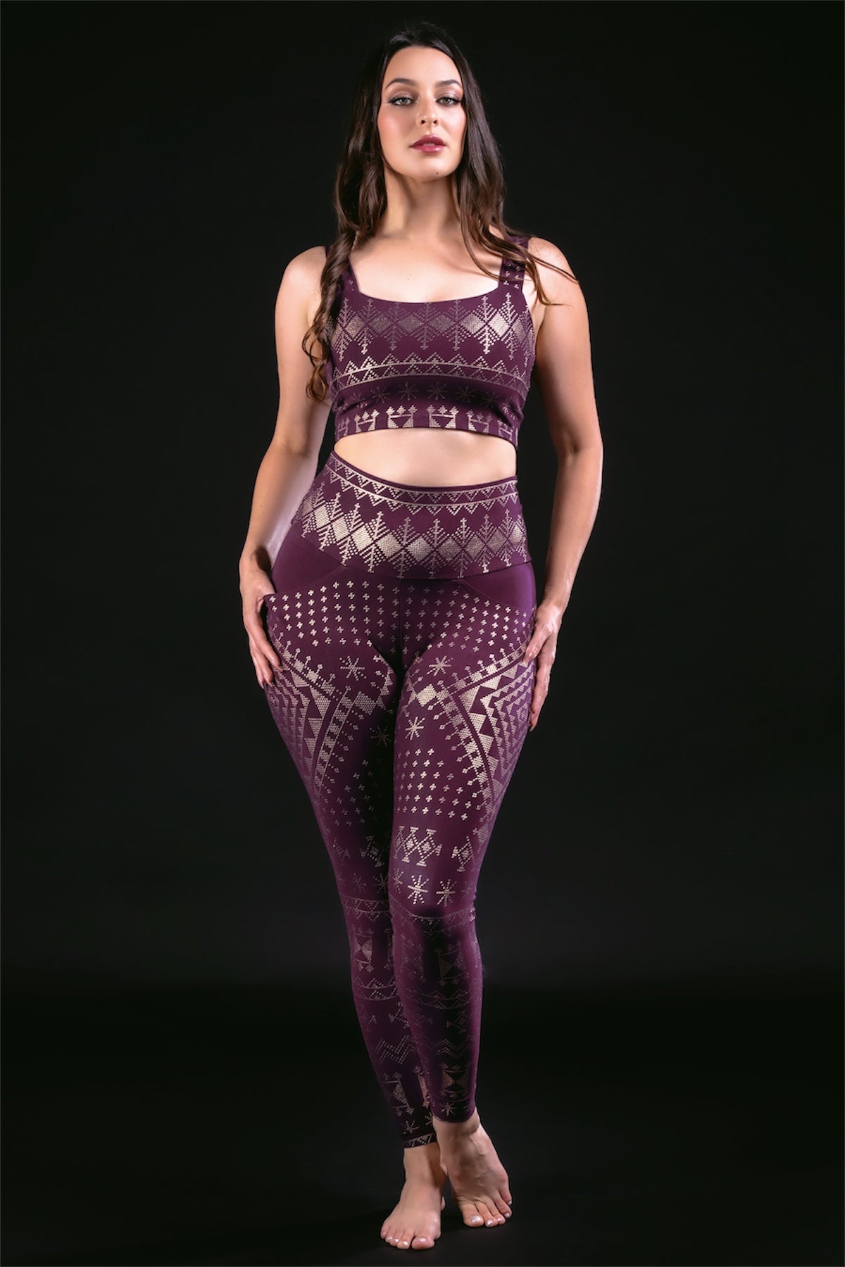 A woman stands barefoot against a black background, wearing Melodia Designs Alkimiya Elsa Legging in Aubergine-Gold with a matching sports bra. The full-length leggings feature gold geometric and snowflake patterns and a comfortable high-rise waistband.