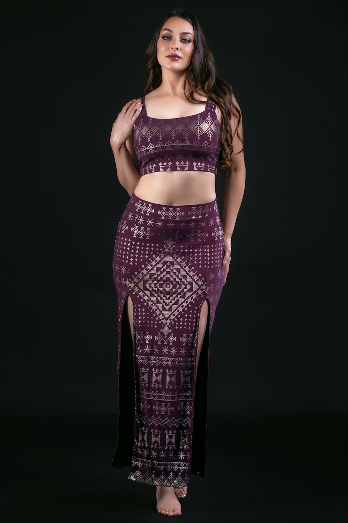 A woman with long brown hair poses confidently barefoot against a black background, wearing the Melodia Designs Alkimiya Neisha Bra in Aubergine-Gold and a matching long skirt with geometric and star motifs and thigh-high slits.