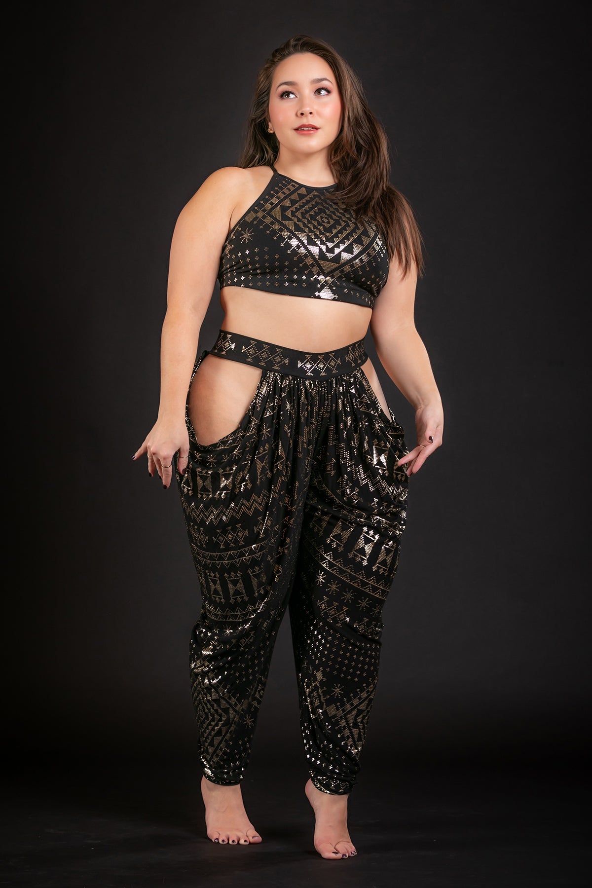A woman stands barefoot against a black background, wearing a shiny black and gold geometric-patterned crop top and Melodia Designs’ Alkimiya Isha Harem Pant Obsidian with large side cutouts, looking slightly to the side with one hand on her hip.