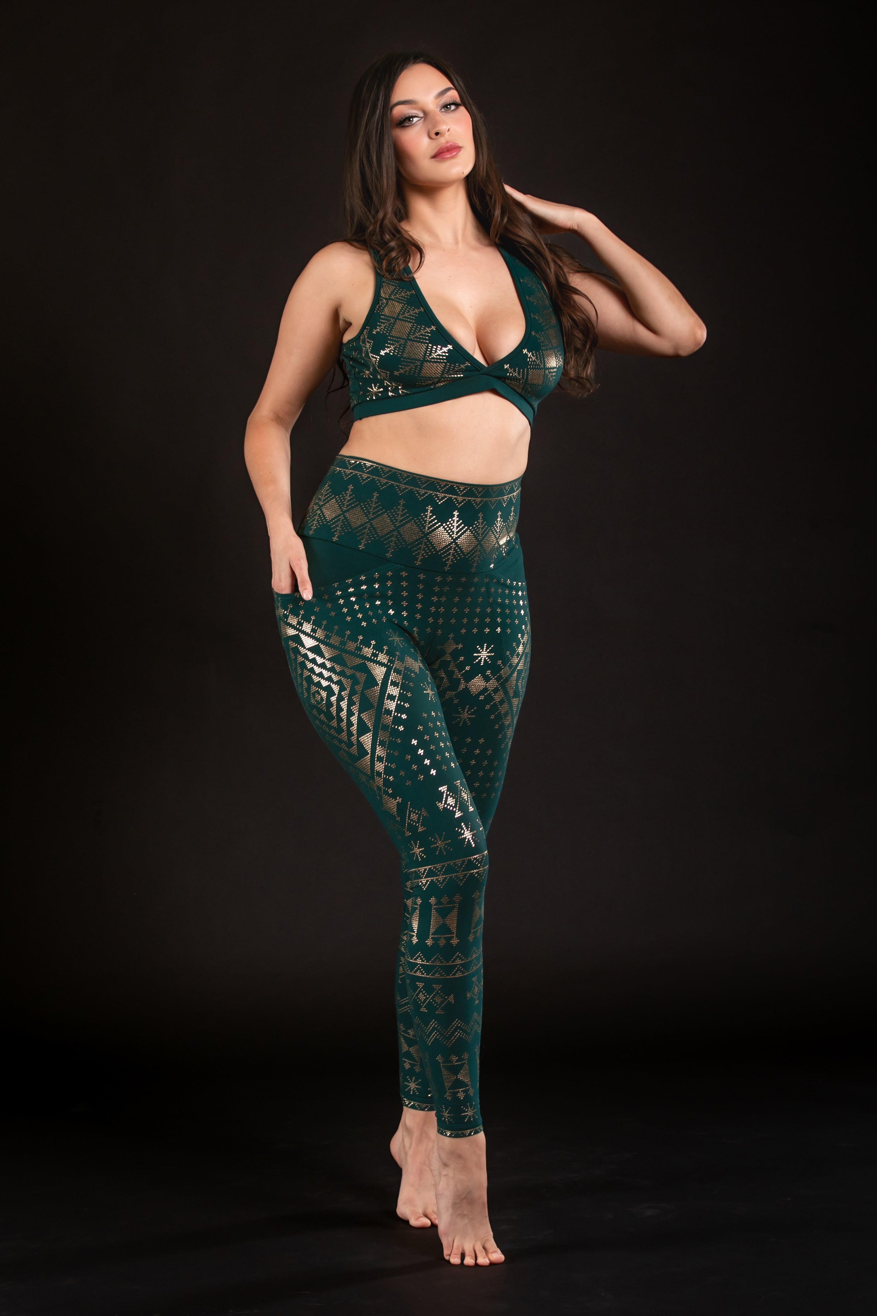 A woman poses barefoot against a black background, wearing the eco-friendly Alkimiya Rainah Bra Malachite by Melodia Designs with matching leggings. She stands with one hand on her hip and the other touching her hair.