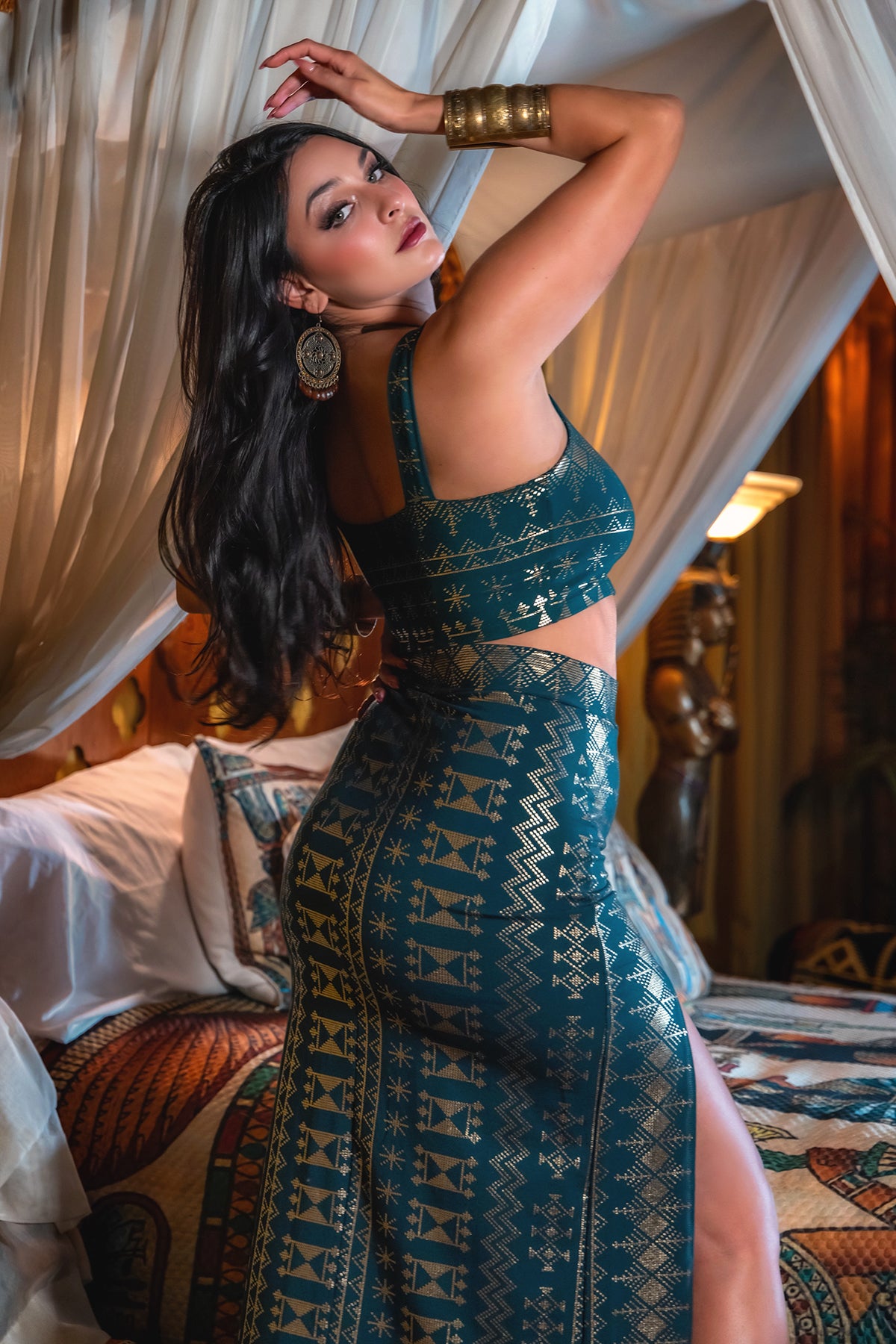 A woman with long dark hair poses indoors by a canopy bed, wearing the Alkimiya Vanessa Skirt Malachite-Gold from Melodia Designs—a sleeveless, patterned green and gold crop top and high-slit skirt made of bamboo organic cotton, accessorized with bold jewelry.