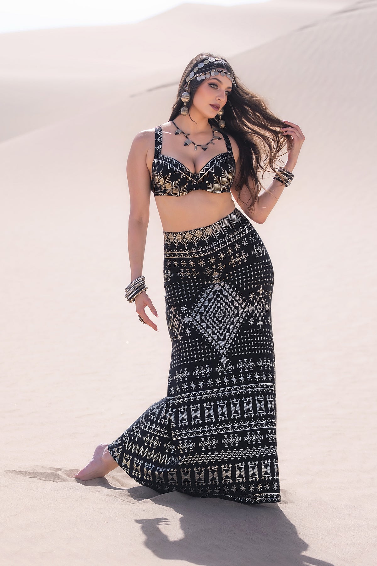 A woman poses barefoot in sand dunes wearing the Melodia Designs Alkimiya Cleopatra Maxi Skirt Obsidian, paired with a matching black and white geometric-patterned crop top, accessorized with jewelry and a headpiece, her hair blowing in the wind.