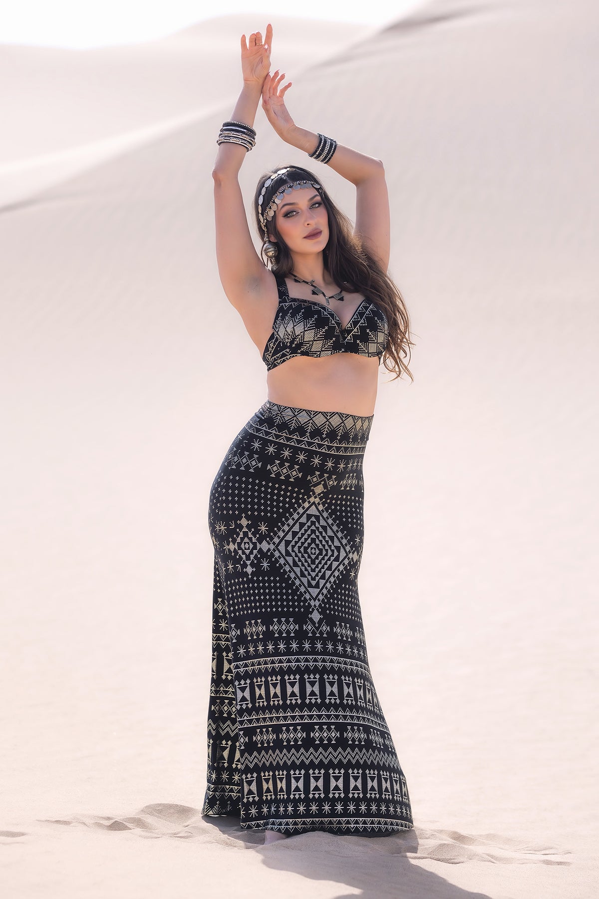 A woman stands in the desert wearing the Melodia Designs Alkimiya Cleopatra Maxi Skirt Obsidian and a black and white crop top. She raises her arms, with long hair, bracelets, a headband, and gazes at the camera against pale sand dunes.