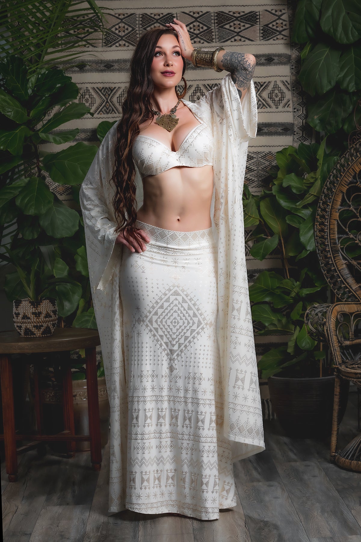 A woman poses indoors in the Melodia Designs Alkimiya Zsa Zsa Robe Ivory, styled with a long patterned white skirt and matching bralette. Lush green plants and woven decor create a glamorous Hollywood-inspired scene.