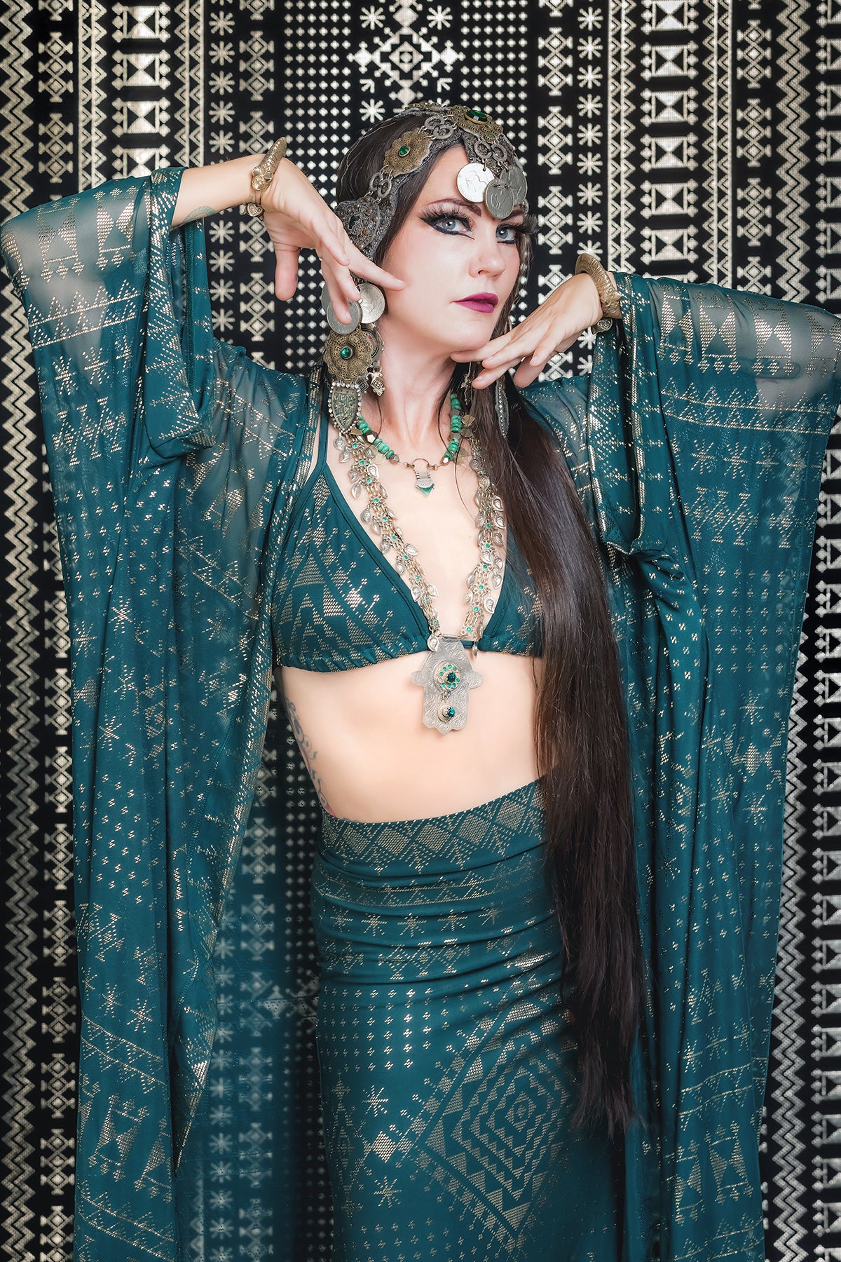 A woman exudes old Hollywood glamour in the Melodia Designs Alkimiya Zsa Zsa Robe Malachite, an elaborate teal and gold statement piece, paired with a striking headpiece and dramatic makeup against a black-and-white patterned backdrop.