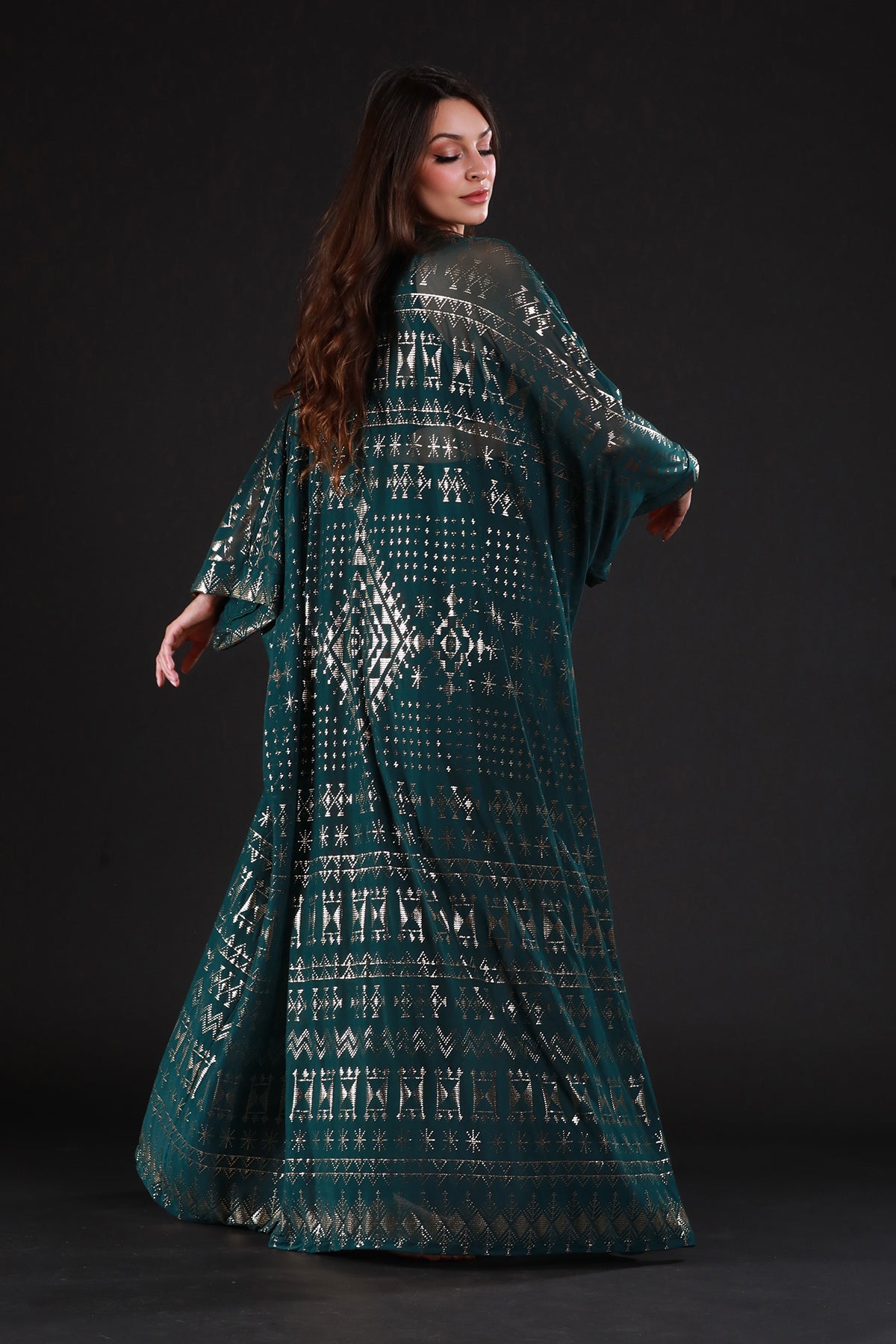 A woman with long brown hair poses gracefully against a dark background, wearing the Alkimiya Zsa Zsa Robe Malachite by Melodia Designs—a flowing dark green dress with gold geometric patterns that channels old Hollywood glamour.