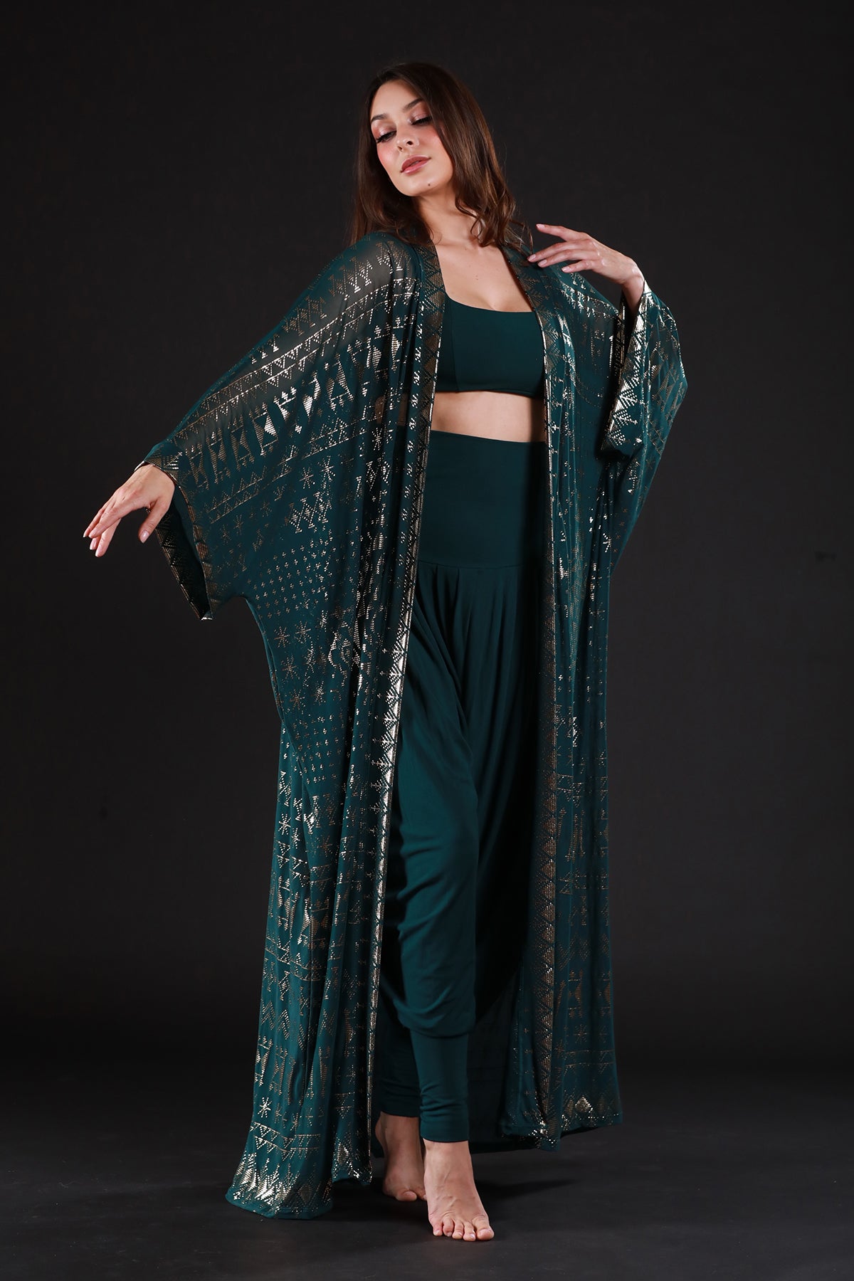 A woman poses barefoot against a black background in the Melodia Designs Alkimiya Zsa Zsa Robe Malachite over a crop top and loose pants, raising one arm gracefully and channeling old Hollywood glamour as she looks to the side.