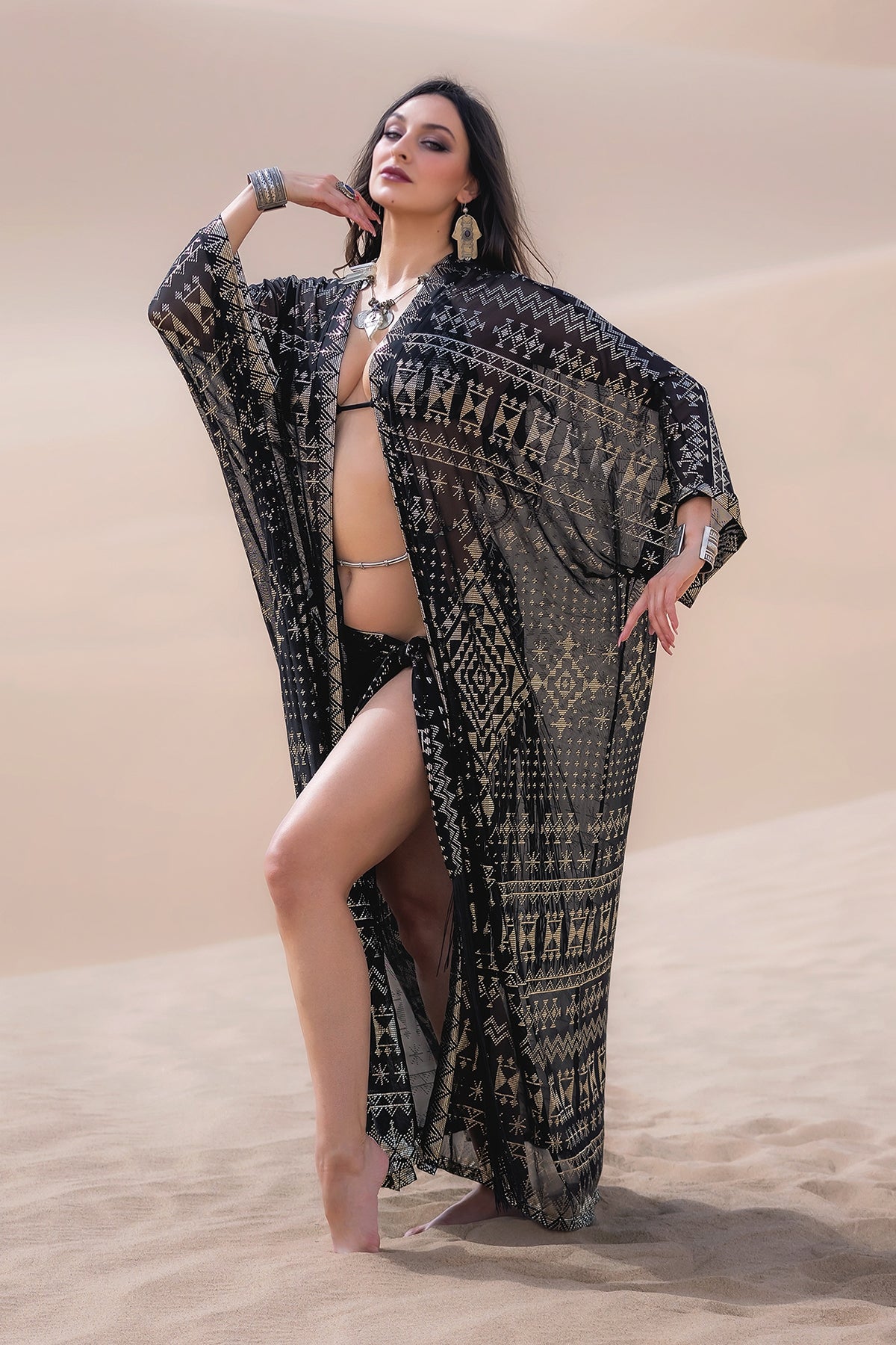 A woman stands barefoot on desert sands in the Melodia Designs Alkimiya Zsa Zsa Robe Obsidian over a bikini, channeling Hollywood glamour with bold earrings, bangles, and a necklace, exuding confidence.