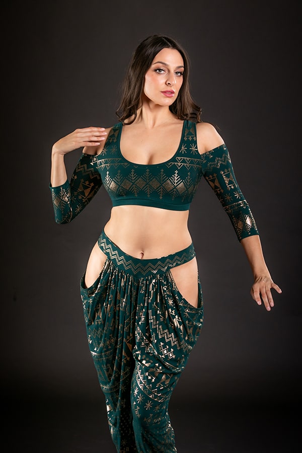 A woman poses gracefully against a black background in the Melodia Designs Alkimiya Amira Cold Shoulder Choli Malachite, featuring stylish cut-out shoulders and hip details in a chic dark green and gold patterned ensemble.