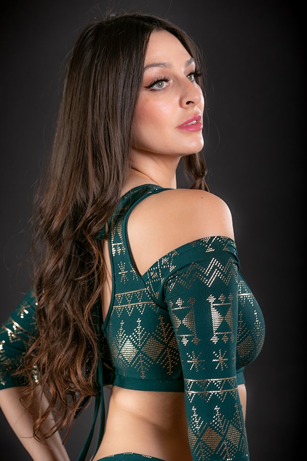 A woman with long brown hair poses against a dark background, wearing the Melodia Designs Alkimiya Amira Cold Shoulder Choli Malachite with geometric patterns and an adjustable back tie, looking over her shoulder.