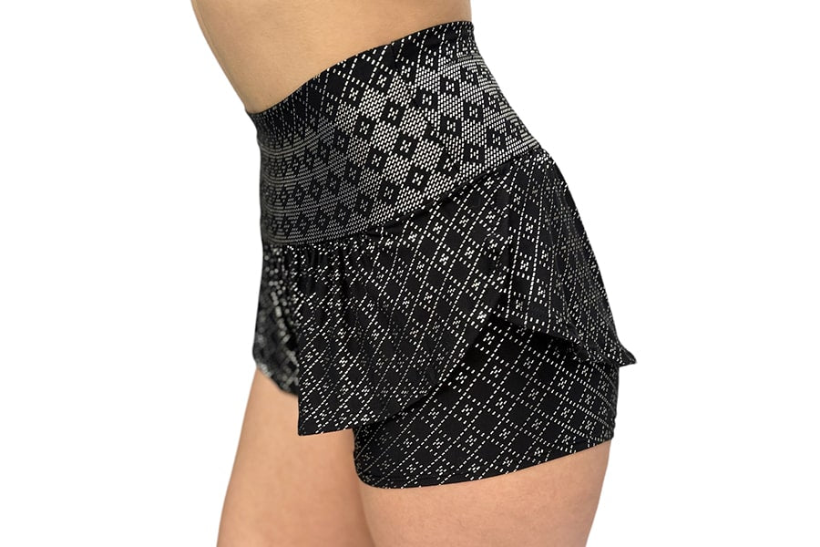 A person in Melodia Designs’ Angela Ruffle Short, featuring a high-waisted, elastic-free fit and black fabric with a white geometric pattern and flared peplum overlay, shown from the side on a white background.
