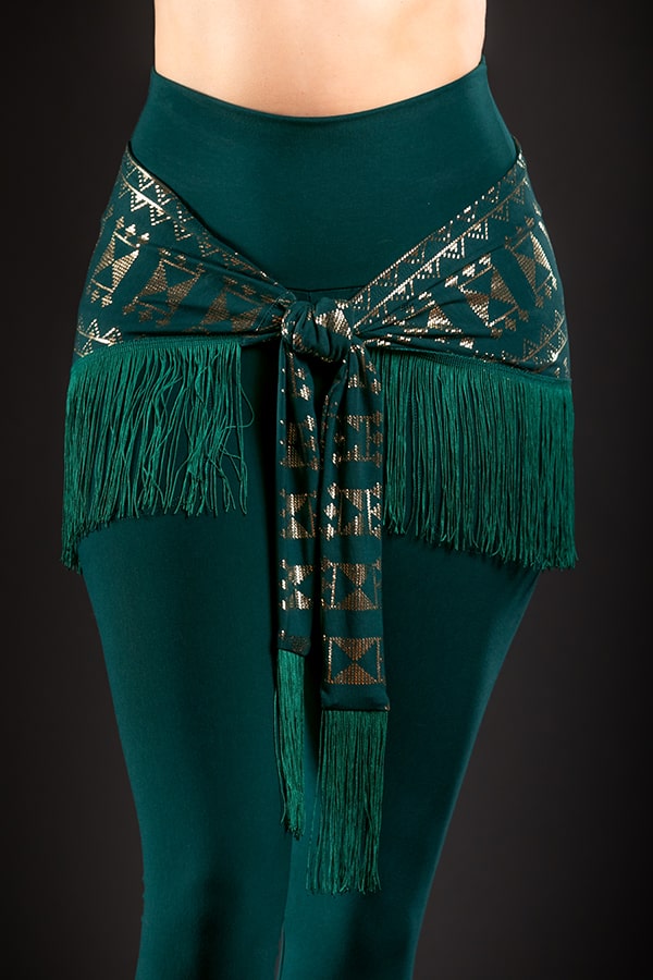 A person wears high-waisted dark green leggings with the Alkimiya Shimmini Shawl Malachite by Melodia Designs, featuring a metallic-patterned chainette fringe, tied at the waist. The black background enhances this striking resort wear look.