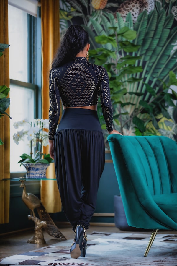 A woman with long dark hair wears the Melodia Designs Auberon Mock Crop Luxor with loose black pants, standing by a green velvet chair in a chic room featuring tropical wallpaper and gold curtains.