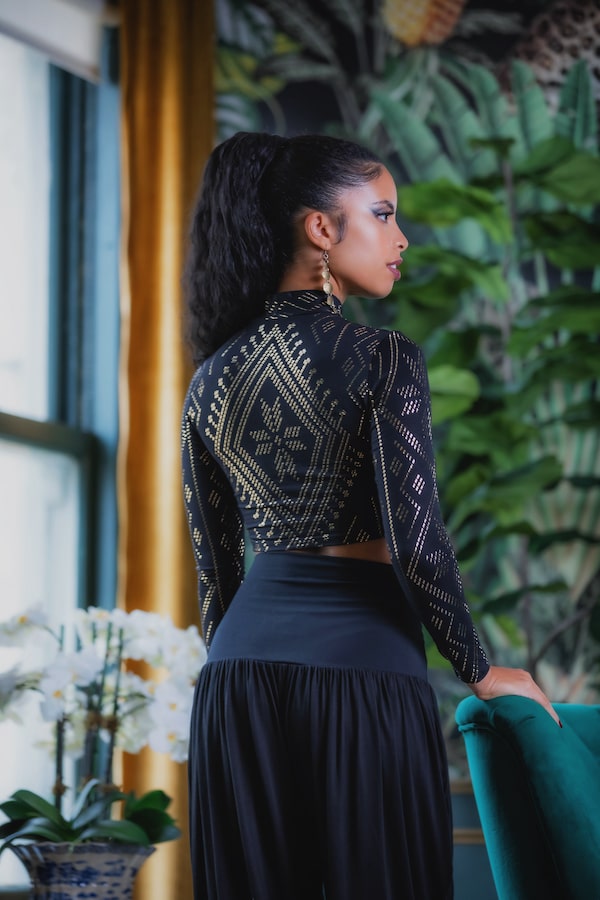 A woman with long curly hair wears the Melodia Designs Auberon Mock Crop Luxor—a black, long-sleeve crop top with gold geometric patterns—paired with a flowing black skirt. She stands indoors near lush green plants and a window.