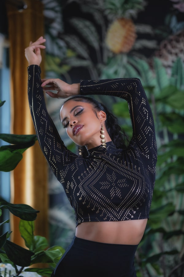 Wearing the Auberon Mock Crop Luxor by Melodia Designs, a woman strikes a graceful pose with arms raised and eyes closed amid lush green plants, large earrings, and a vibrant tropical-themed backdrop.