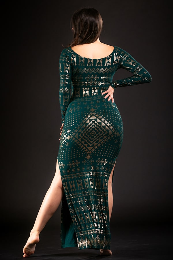 A woman stands barefoot on tiptoe, facing away, wearing the Melodia Designs Alkimiya Nawal Dress Malachite—long, dark green with gold geometric patterns and side slits—set against a dark background.