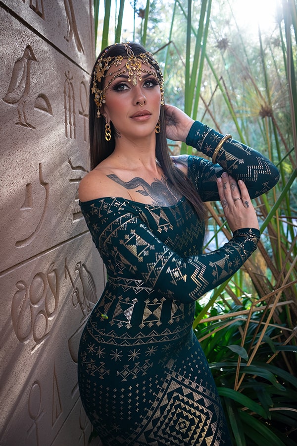 A woman wears the Alkimiya Nawal Dress Malachite by Melodia Designs, adorned with gold jewelry beside a wall of Egyptian hieroglyphs. She stands among lush green plants in gentle sunlight.