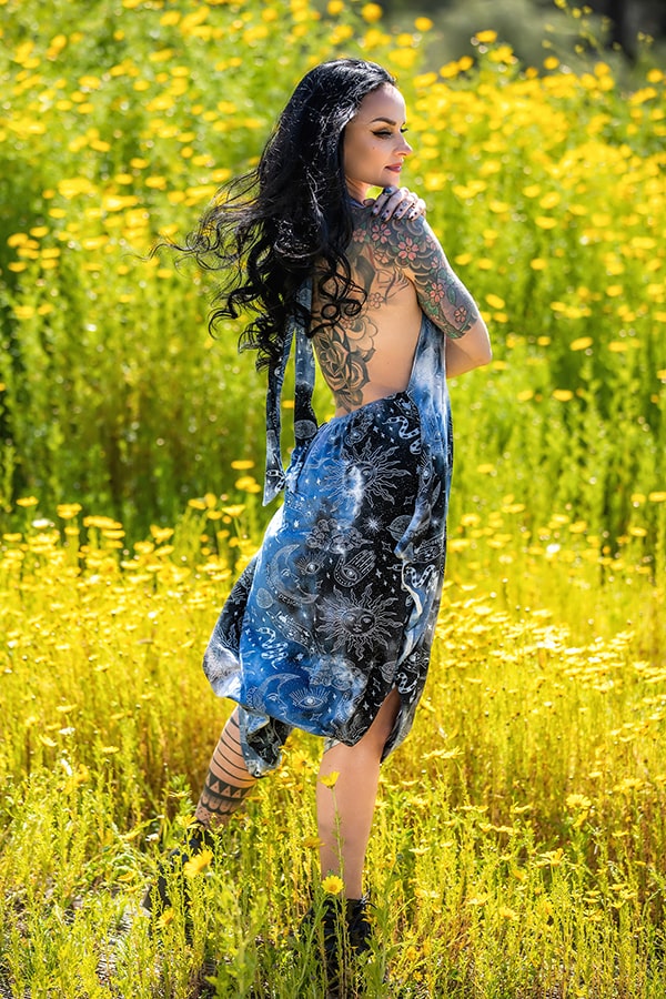 A woman with long dark hair and tattoos stands in a field of yellow wildflowers, smiling over her shoulder in the Melodia Designs Briana Jumper Tie Dye Celestial, which features a backless celestial-inspired print.