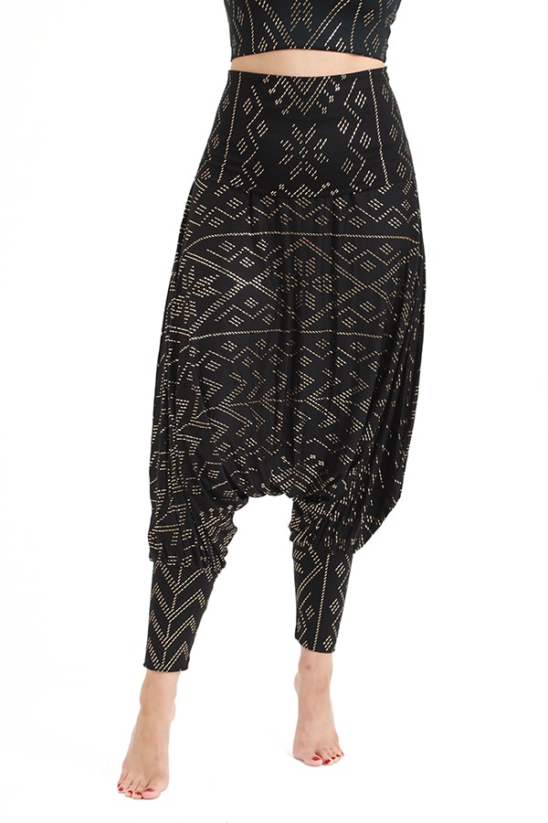 A person models the Christine Luxor Harem Jumper by Melodia Designs—high-waisted, loose black pants with tan geometric patterns, tapered ankles, and a dropped crotch—styled barefoot against a white background.