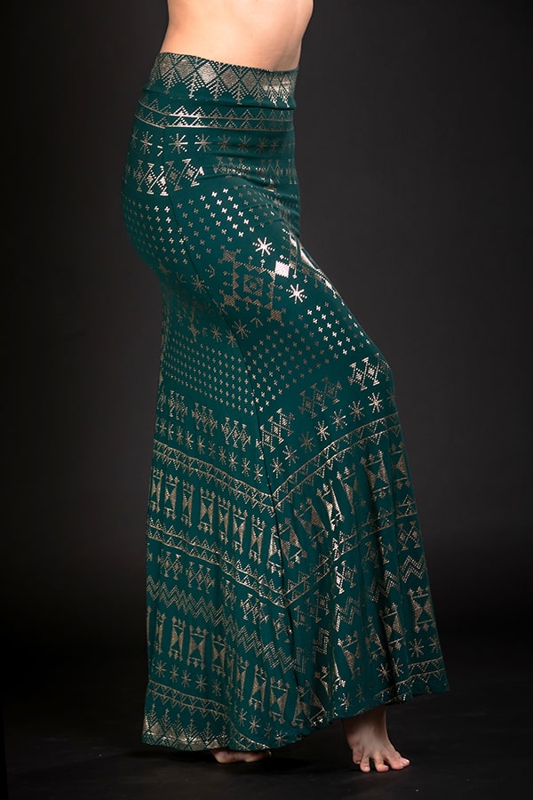 A person stands barefoot in a Melodia Designs Alkimiya Cleopatra Maxi Skirt Malachite—a long, dark green A-line skirt with gold geometric and tribal patterns—posing against a plain black background. Only the lower torso and legs are visible.