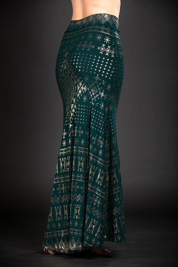 A person wears the Alkimiya Cleopatra Maxi Skirt Malachite by Melodia Designs—an A-line, high-waisted teal skirt with gold geometric and star patterns—standing barefoot against a dark background.