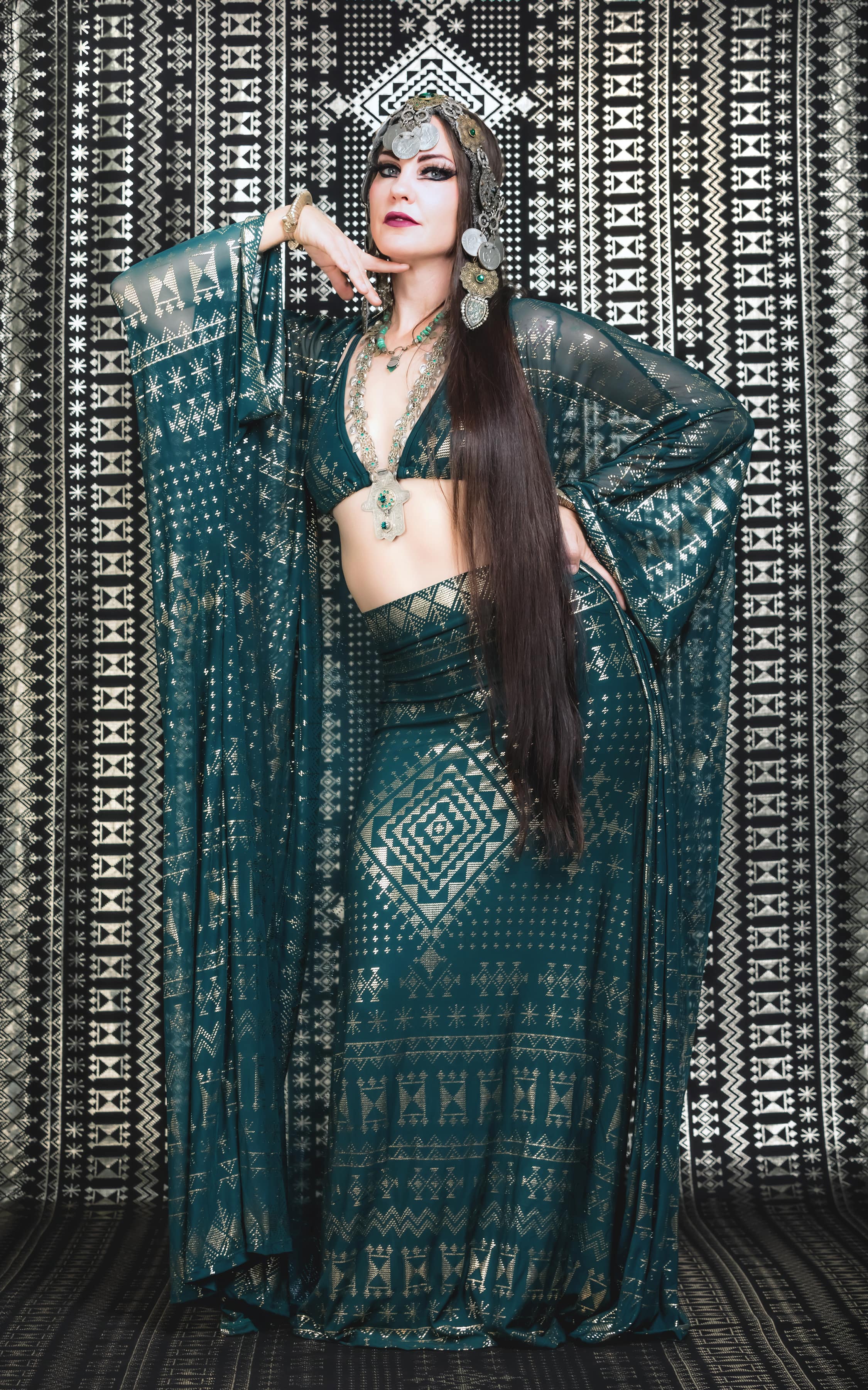 A woman models the Alkimiya Cleopatra Maxi Skirt Malachite by Melodia Designs, styled with an ornate headdress, statement jewelry, and long dark hair, posing gracefully before a black-and-white patterned backdrop.