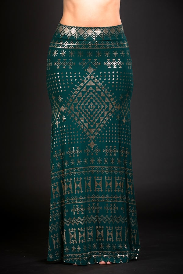 A person stands against a black background wearing the Melodia Designs Alkimiya Cleopatra Maxi Skirt Malachite, an A-line, floor-length skirt in dark green with gold geometric and tribal patterns, featuring a fitted waist and graceful flow.