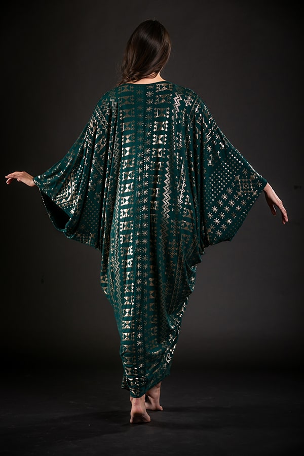 A woman stands barefoot with her back to the camera, wearing the Alkimiya Contessa Caftan Malachite by Melodia Designs. The eco-luxe fabric’s green and gold pattern and wide batwing sleeves are displayed against a dark background.