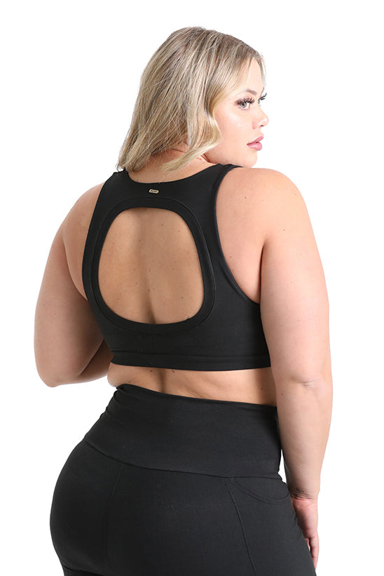 A blonde woman wears the Melodia Designs Serena Bra 2.0, a black eco-luxe supportive bra with a large oval back cutout, paired with matching high-waisted leggings, posing with her back to the camera against a white background.