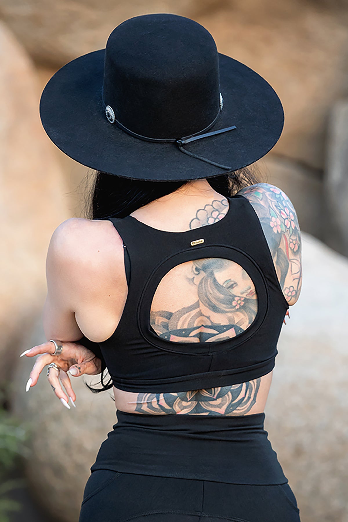 A woman with long dark hair and colorful tattoos wears the Melodia Designs Serena Bra 2.0, made from eco-luxe fabric, and a black wide-brimmed hat as she stands outdoors facing away from the camera.