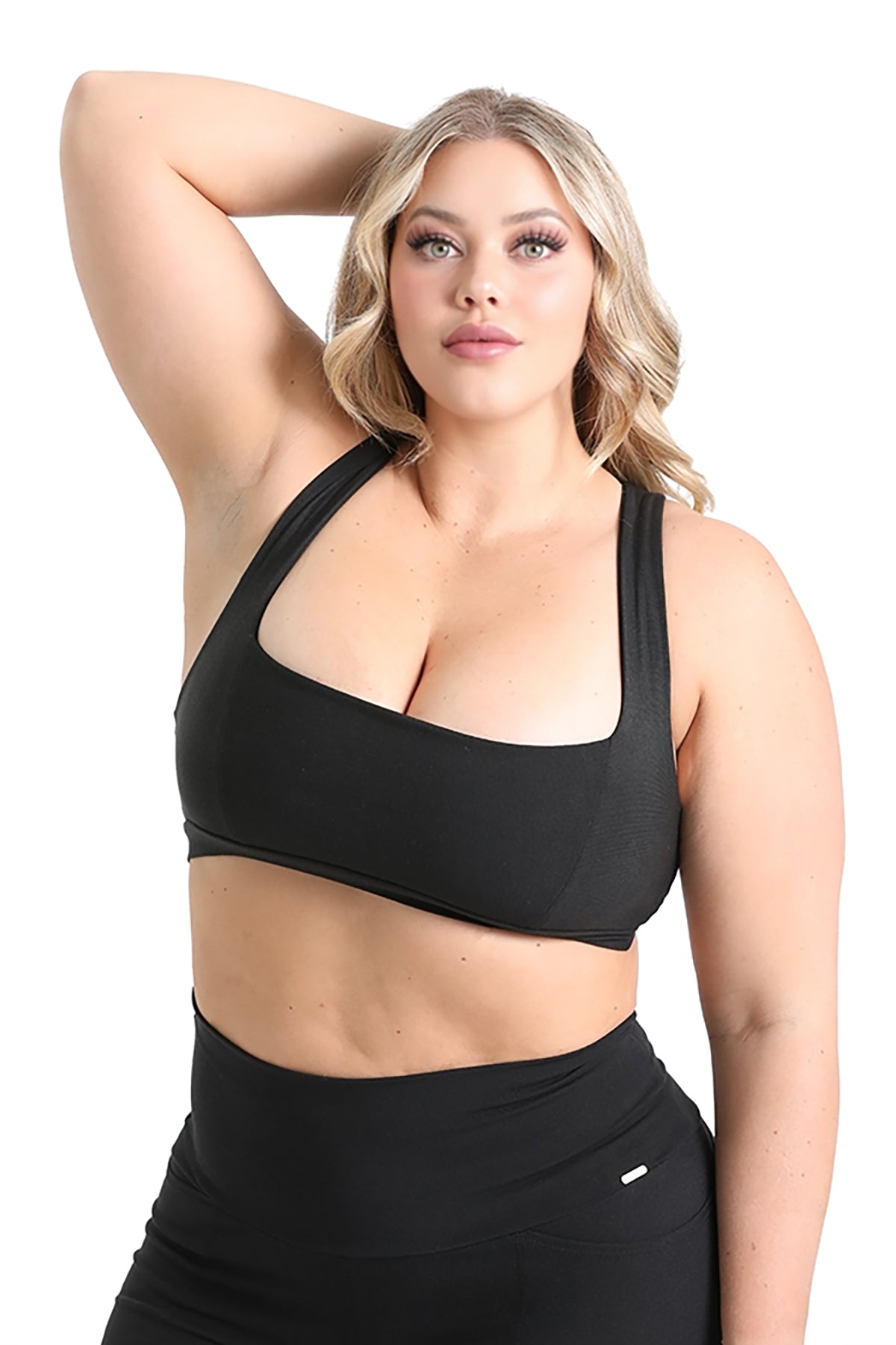 A woman with long, wavy blonde hair models the Melodia Designs Serena Bra 2.0 in black with matching high-waisted leggings, posing confidently with one arm raised behind her head against a plain white background.