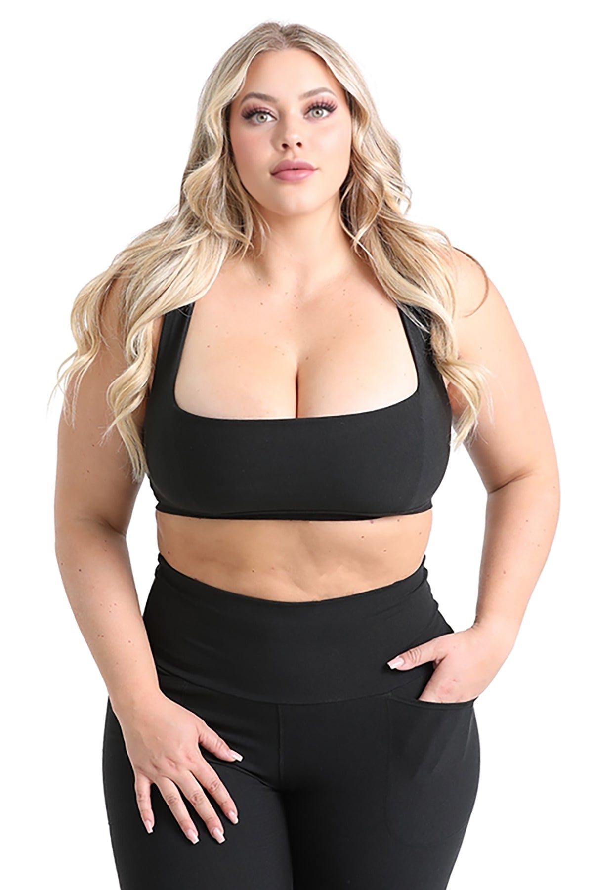 A woman with long blonde hair models the Melodia Designs Serena Bra 2.0, a black crop top made from eco-luxe fabric, paired with high-waisted black pants, posing confidently with one hand on her hip against a white background.