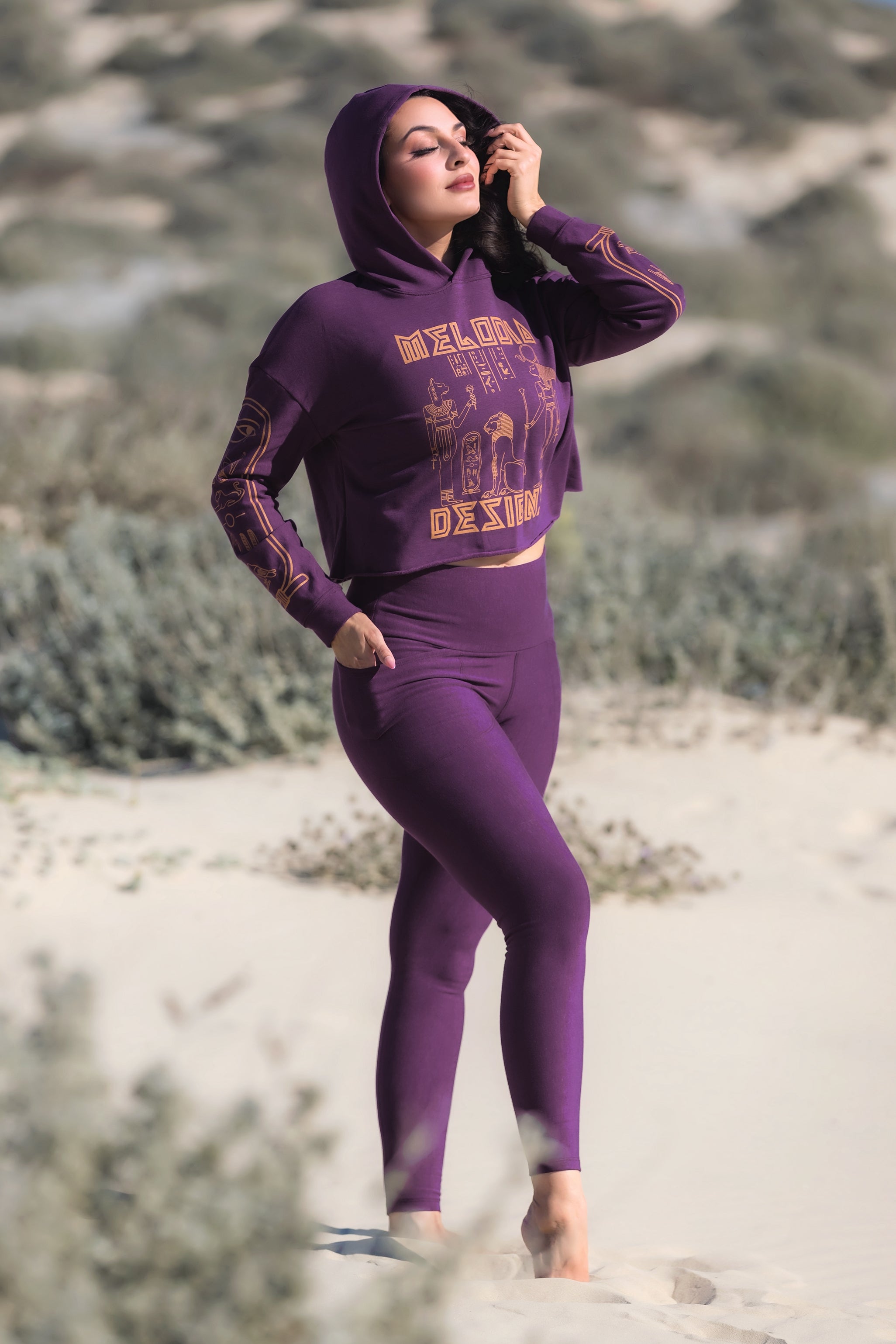 A person wears Melodia Designs’ Elsa Legging Fleece Aubergine with a foldable waistband, standing barefoot on sand. They touch their face and look relaxed, with dunes and sparse vegetation in the background.