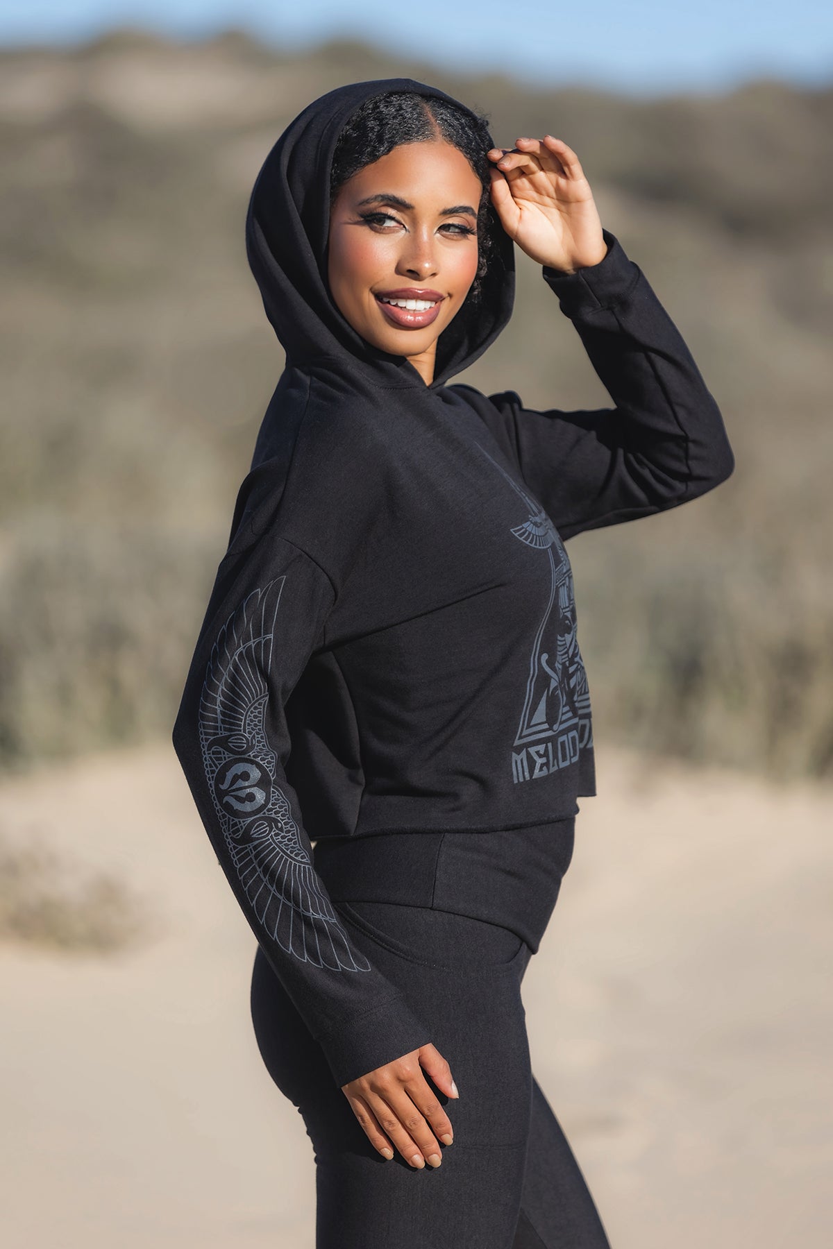 A woman smiles outdoors in the Melodia Designs Crop Hoodie Fleece Black, holding her hood. The cozy bamboo fleece hoodie features detailed wing designs on the sleeves. The matching pants and blurred natural background complete the look.