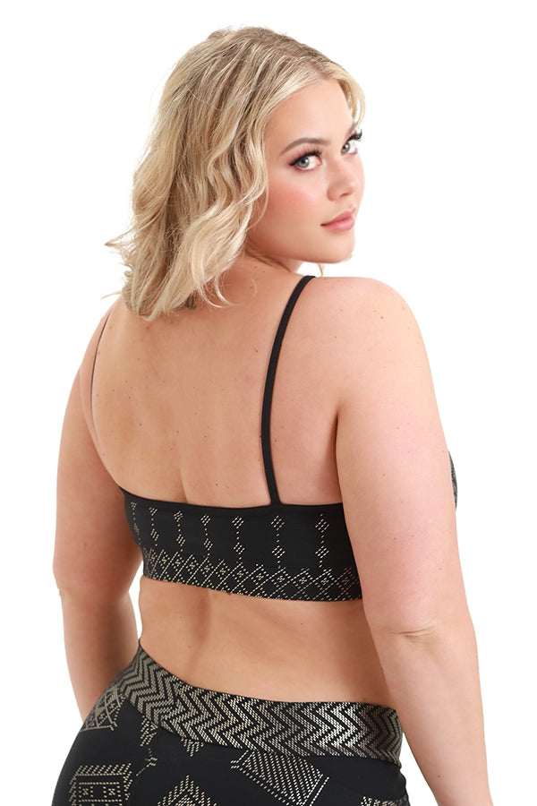 A woman with short blonde hair poses with her back to the camera, wearing the Kora Bra by Melodia Designs—black with gold geometric patterns and matching performance leggings—against a white background.