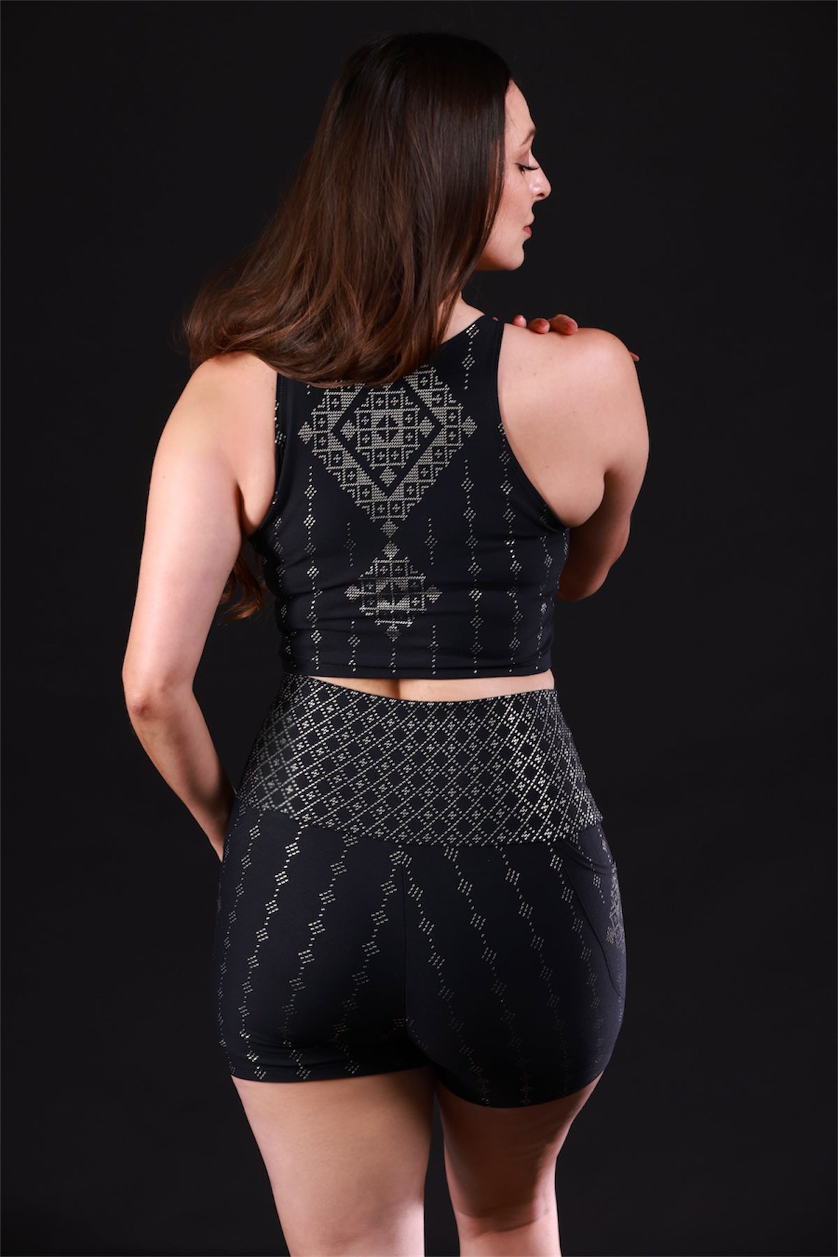 A woman with long brown hair, wearing a black patterned sports bra and Melodia Designs Deco Alice Short Black-Antique Silver, stands facing away from the camera against a solid black background.