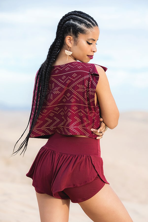 A woman with long braids stands in a desert, wearing Melodia Designs’ Angela Ruffle Short and a sleeveless maroon top with geometric patterns. She looks over her shoulder, highlighting her eco-luxe outfit and elegant earrings.