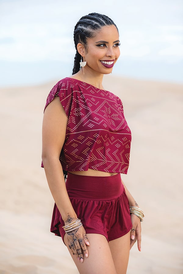 A smiling woman with braided hair stands in a sandy desert wearing Melodia Designs’ burgundy Angela Ruffle Short and a peplum crop top, accessorized with earrings, bracelets, and henna on her left hand.