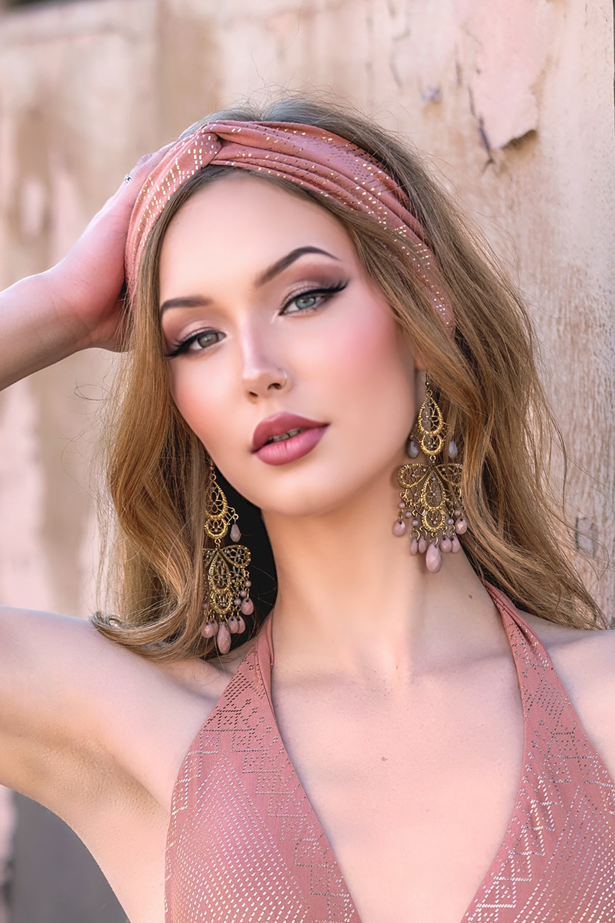 A woman with long, wavy blonde hair wears a pink Melodia Designs Dendera Beauty Band Desert Rose and gold chandelier earrings, posing with one hand on her head by a textured, light-colored wall.