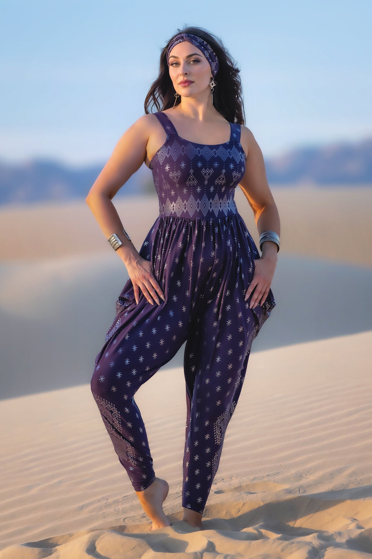 A woman stands barefoot on sand dunes, wearing the Melodia Designs Dendera Jinni Jumper 2.0 Midnight Silver with a matching headband and silver jewelry. She has long dark hair, and mountains are visible in the background.