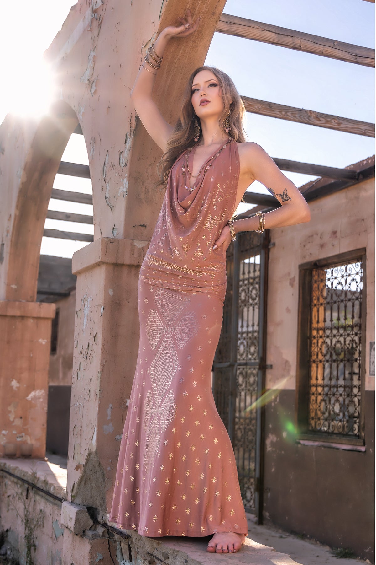 A woman models the Melodia Designs Dendera Raja Halter Desert Rose Gold dress, posing barefoot in sunlight by a weathered stone archway, her arm raised and her gaze uplifted.
