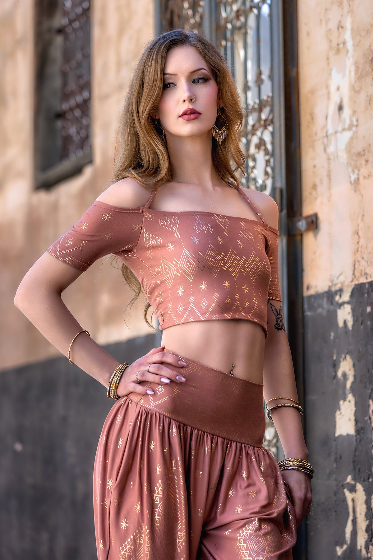 A woman in Melodia Designs' Dendera Zaina Harem 2.0 Desert Rose Gold pant and a peach off-shoulder crop top stands confidently outdoors by a textured wall and decorative window, wearing gold jewelry with long, wavy hair.