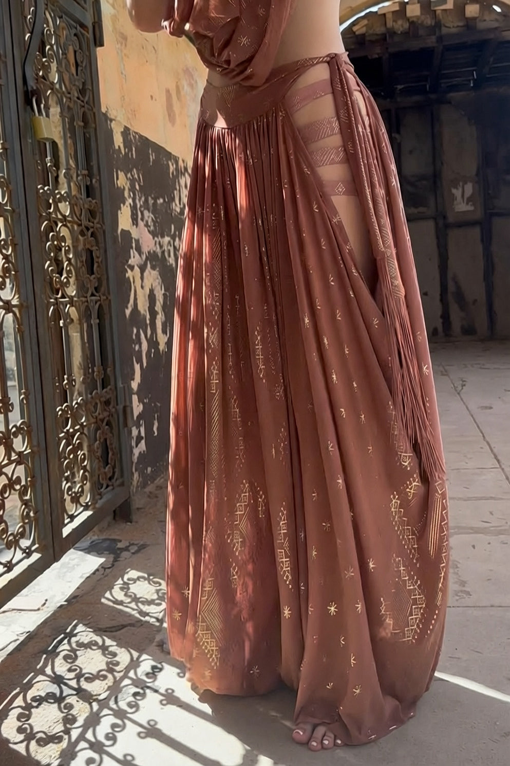 A person wears Melodia Designs’ Dendera Fringe Sahara Pantaloon in Desert Rose, featuring gold geometric accents and side cutouts, standing barefoot on a sunlit stone floor by an ornate metal gate and weathered walls.