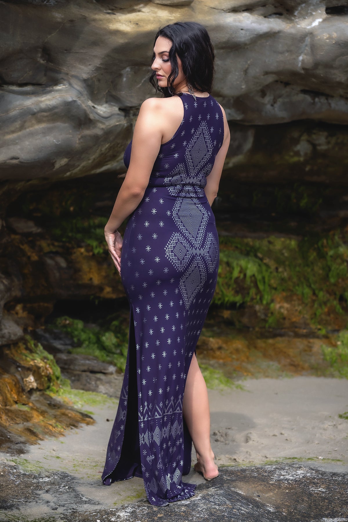 A woman stands outdoors on sandy ground, facing away, wearing the Melodia Designs Dendera Shauntel Dress in Midnight Silver. Rocky formations and green moss patches appear in the background.
