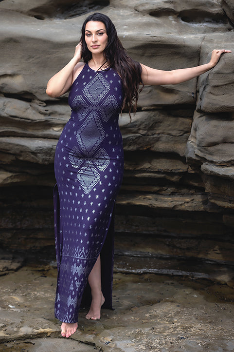 A woman with long dark hair stands barefoot on rocks, confidently posing in the Melodia Designs Dendera Shauntel Dress Midnight Silver, a sleeveless, form-fitting dress with geometric patterns.