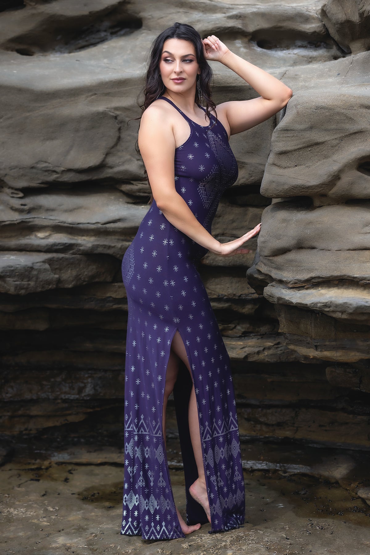 A woman stands barefoot on rocky terrain, confidently gazing ahead in the Melodia Designs Dendera Shauntel Dress Midnight Silver, a sleeveless fitted piece with a geometric pattern and side slit.