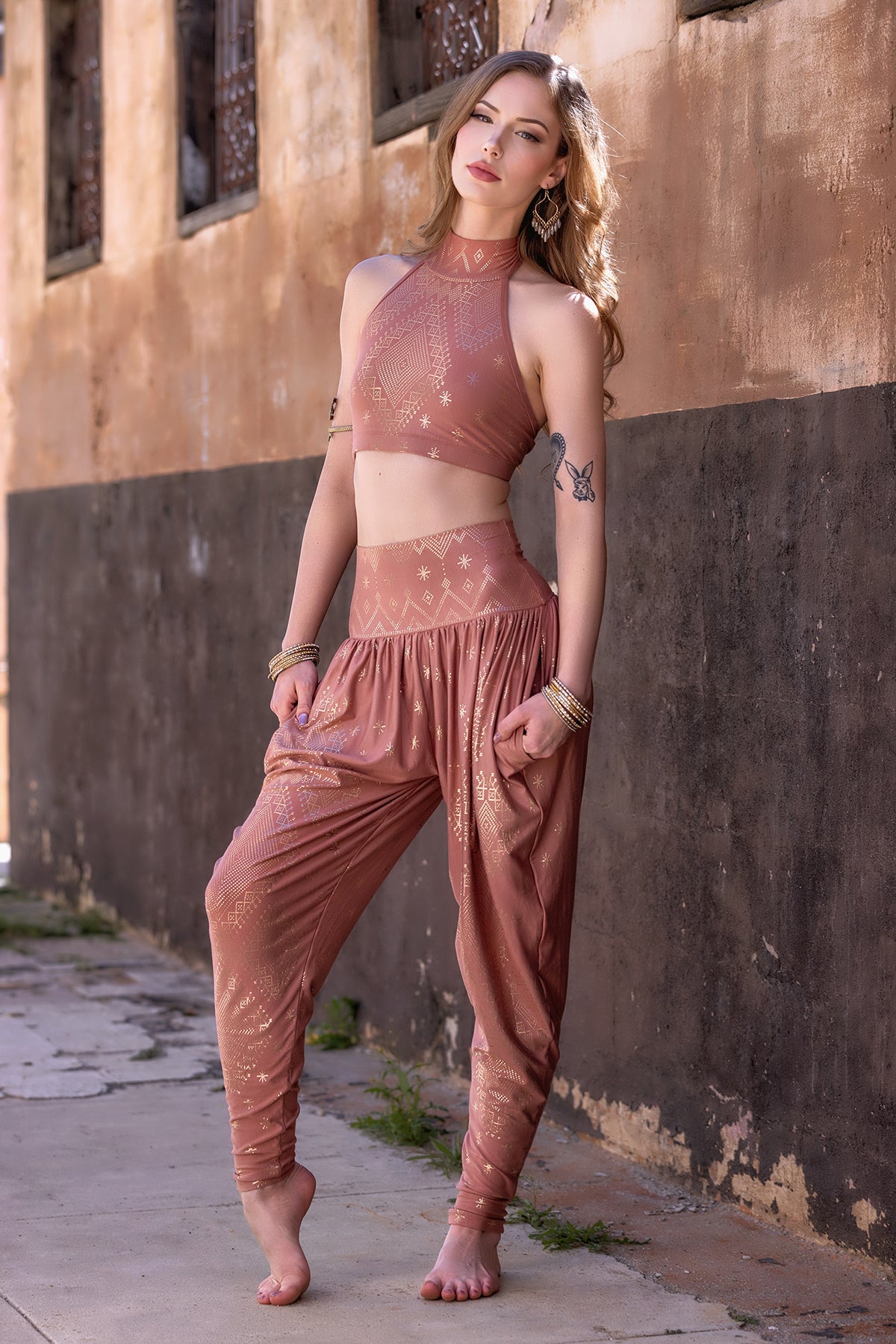 A barefoot woman leans against a worn wall, wearing the Melodia Designs Dendera Zaina Harem 2.0 Desert Rose Gold pants with a matching mauve crop top, accessorized with bracelets, earrings, and a forearm tattoo as she gazes at the camera.
